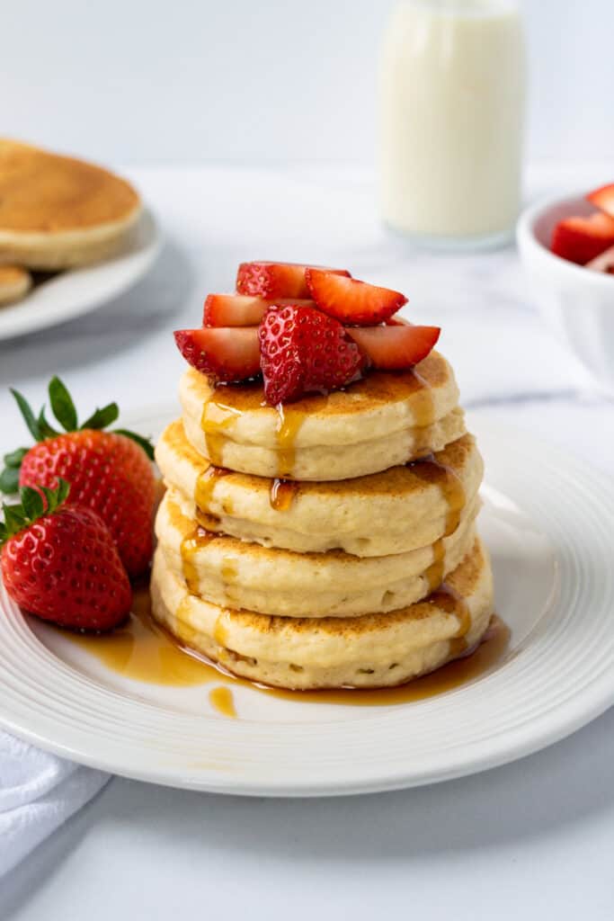 Stack of 4 oat milk pancakes on a plate topped with strawberries and maple syrup.