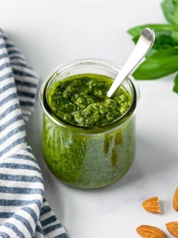 Almond Basil Pesto in a glass jar on a table with almonds scattered around it.