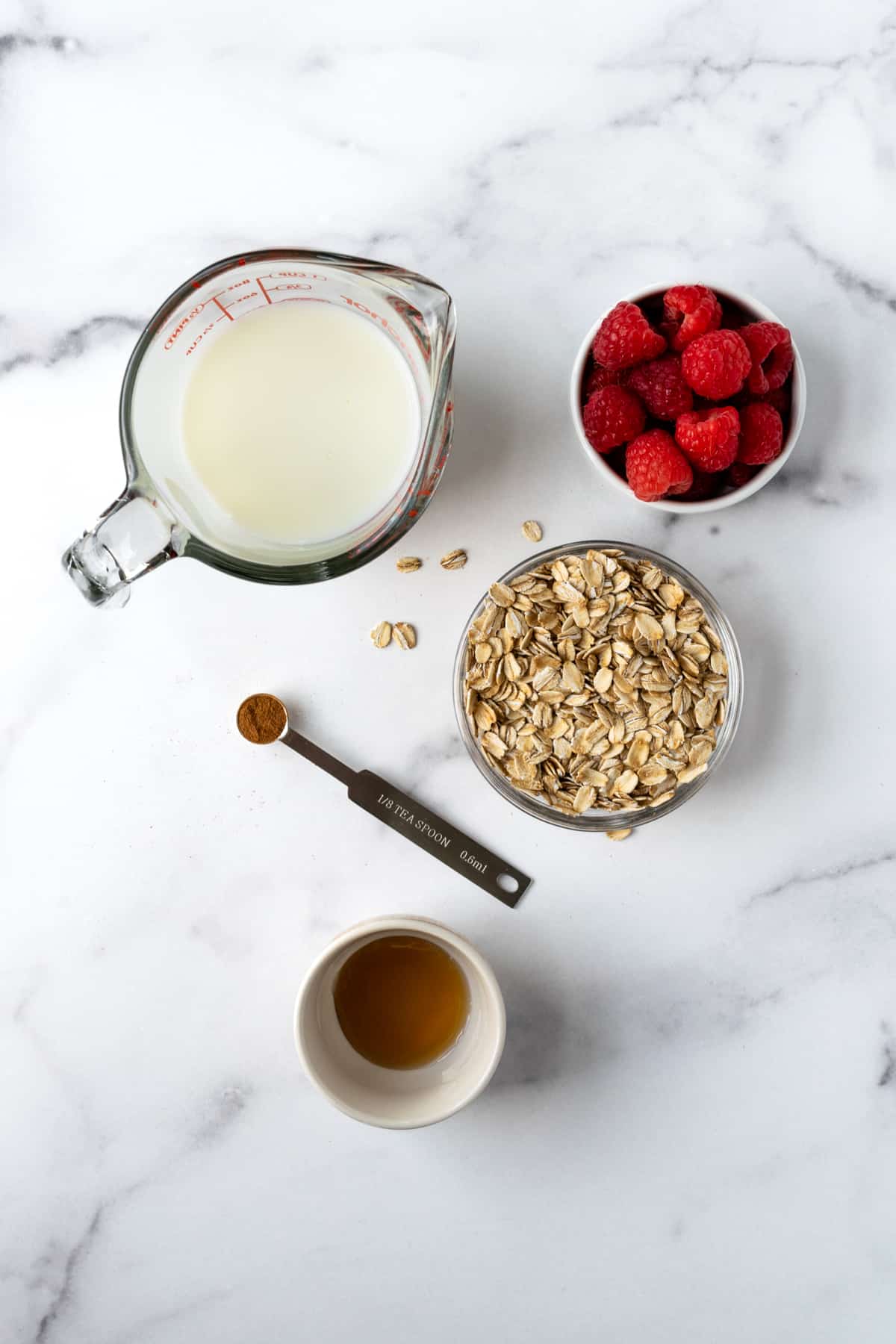Milk, Oats, Raspberries, Cinnamon, and Maple Syrup in prep bowls on a marble countertop for overnight oats.