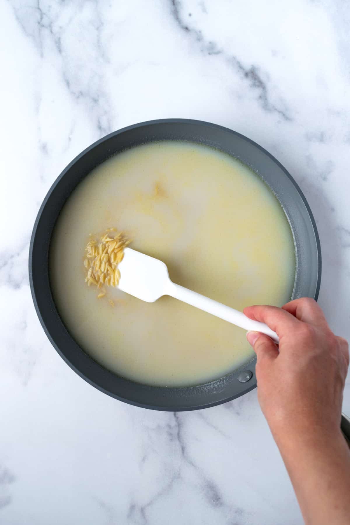 Orzo, broth, and milk in a skillet with a white silicone spatula.