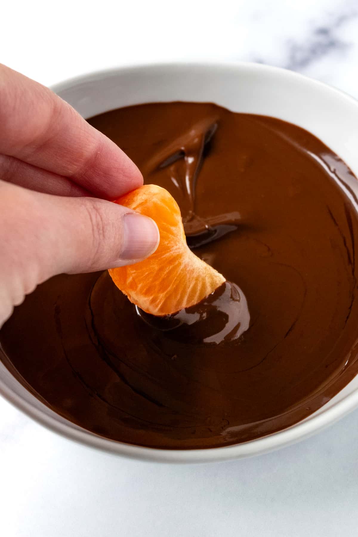 Hand dipping a mandarin orange in melted chocolate.