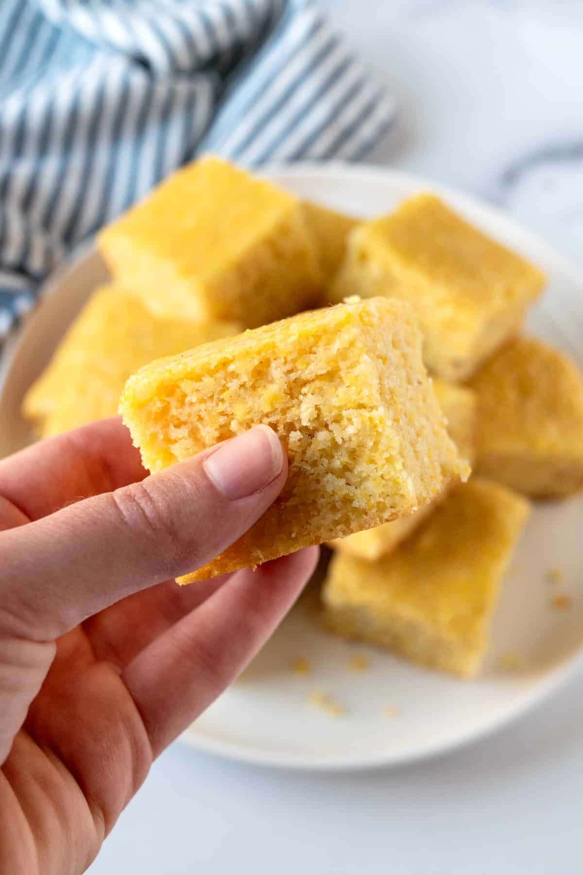 A hand holding a piece of cornbread, with a plate of cornbread in the background.
