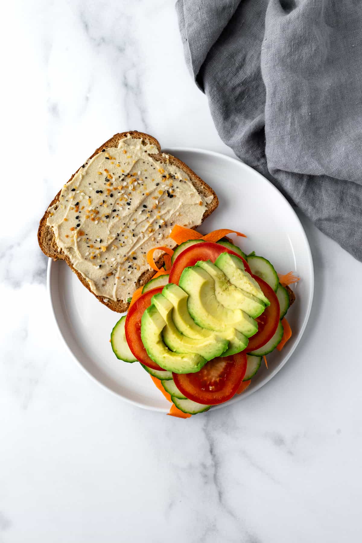 Open faced Cucumber Hummus Sandwich topped with carrots, tomato, and avocado on a white plate.
