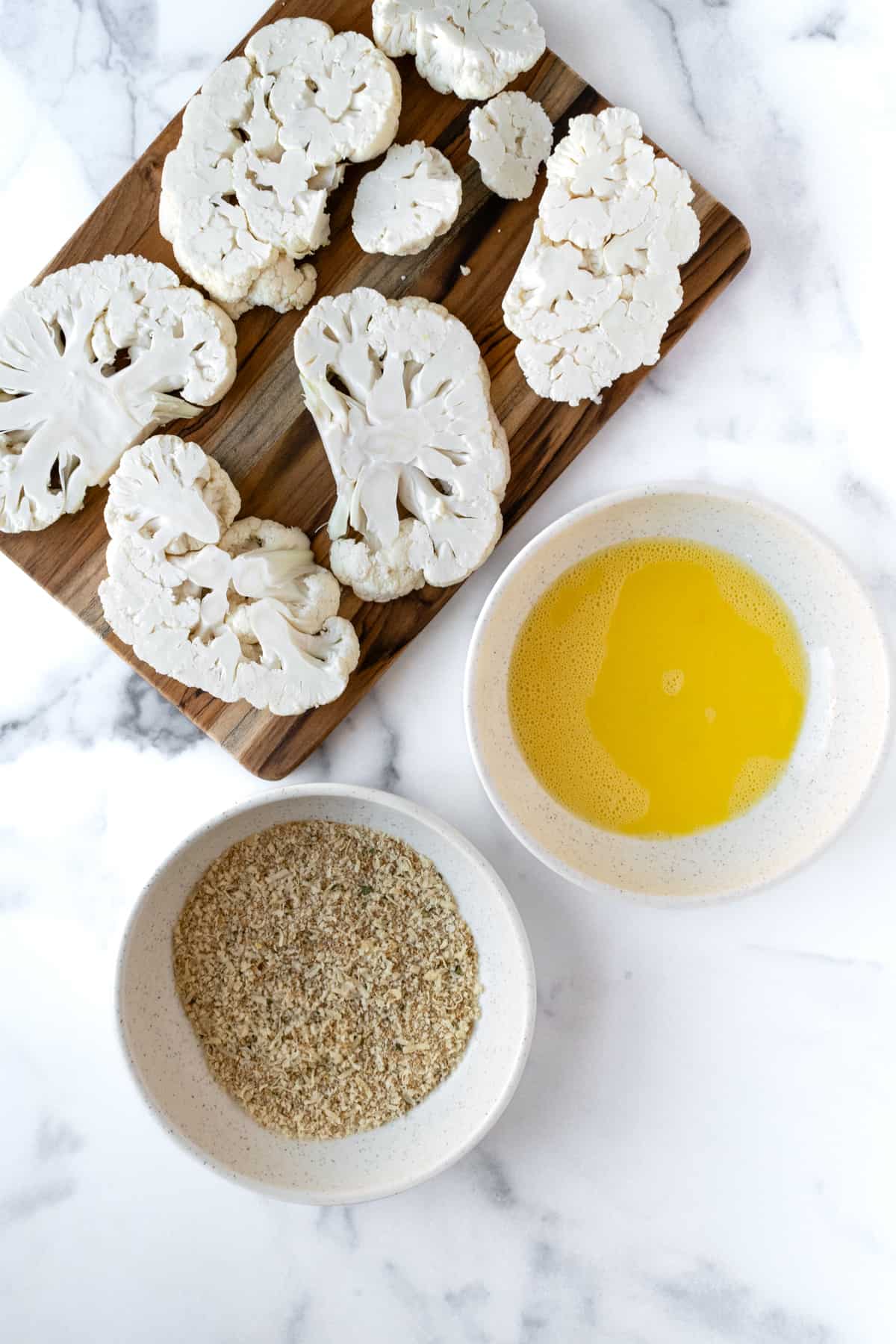 Raw Cauliflower Steaks on a cutting board next to an egg wash and a bowl of breadcrumbs.