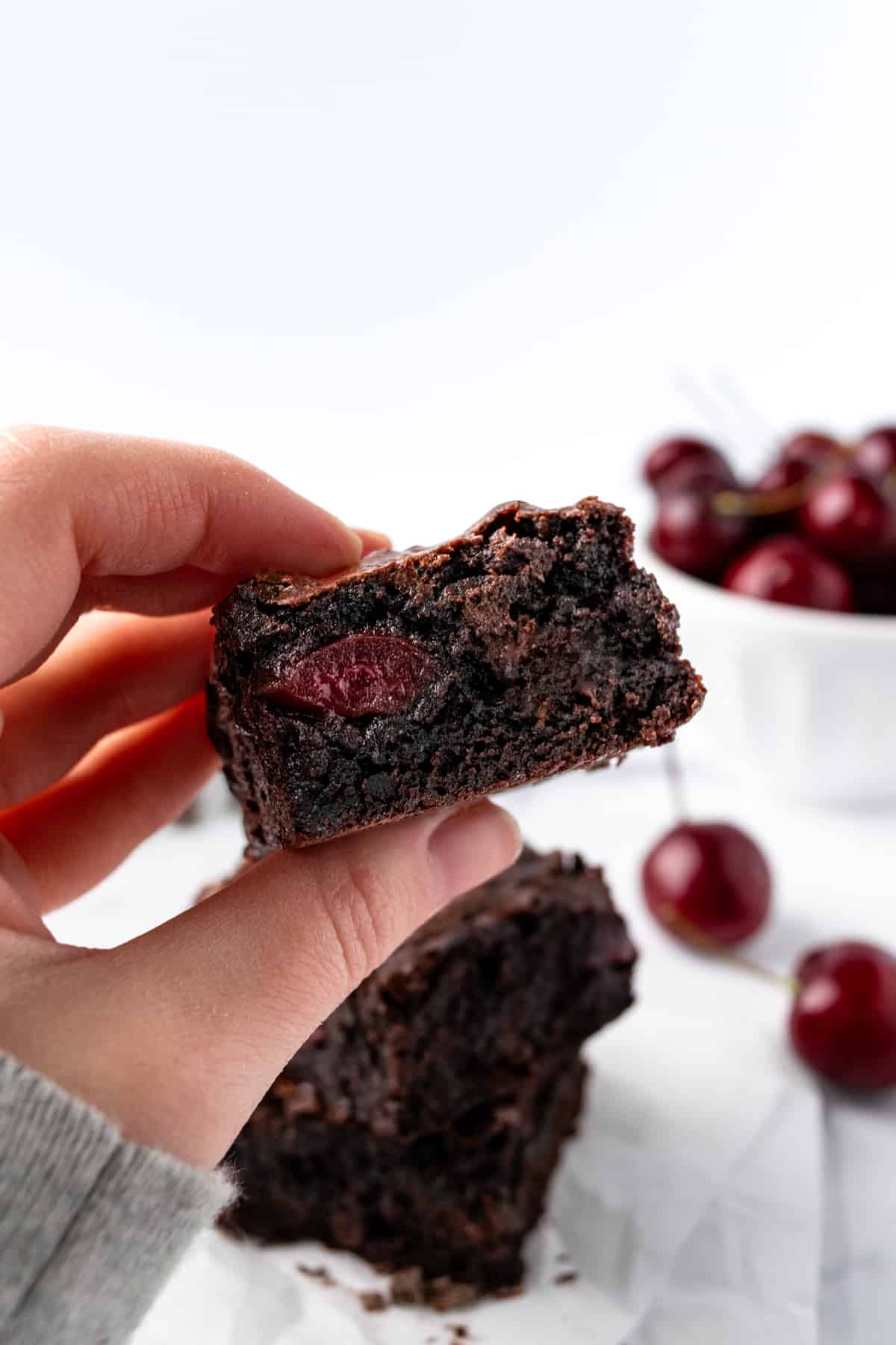 A hand holding a cherry chocolate brownie with a bowl of fresh cherries in the background.