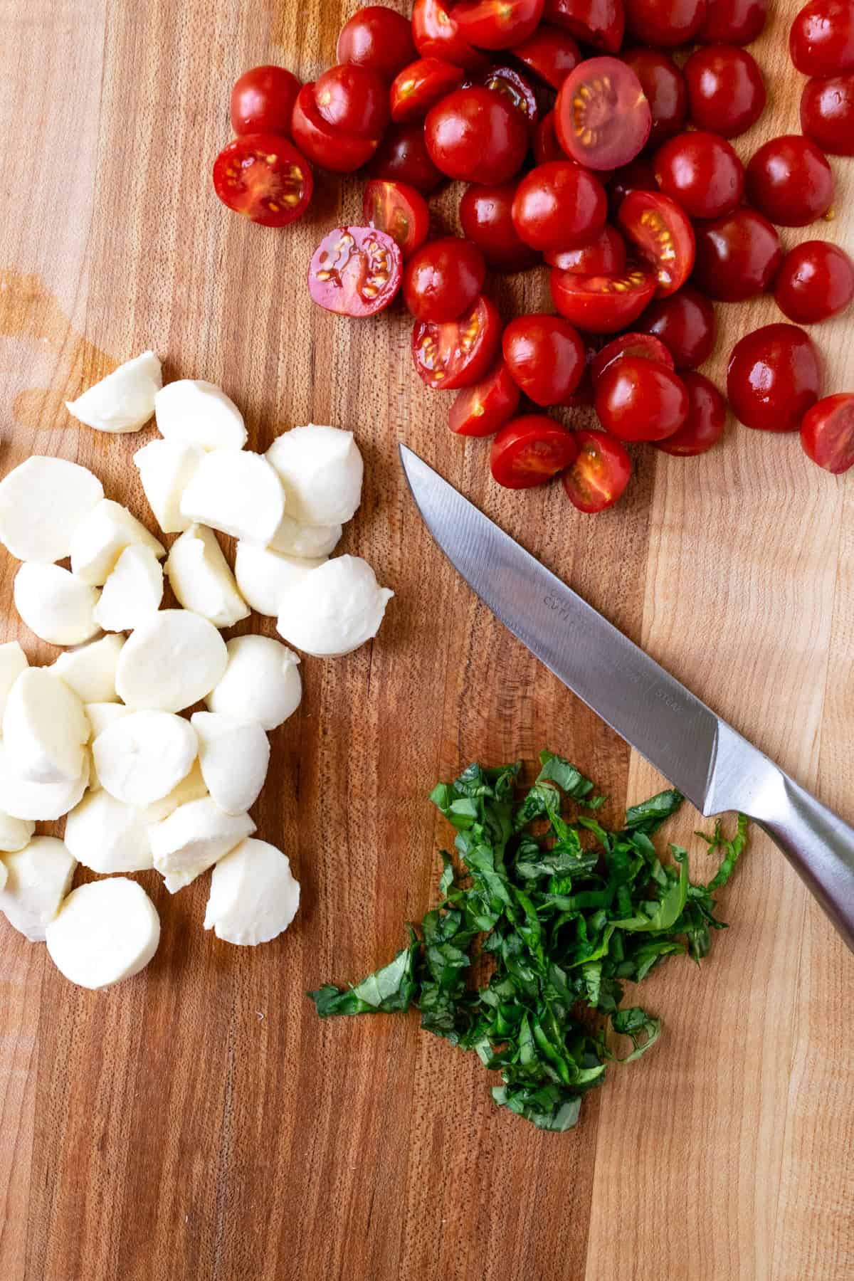 Chopped cherry tomatoes, mozzarella, and fresh basil on a cutting board with a knife.