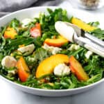 Peach and Pistachio Arugula Salad in a white serving bowl with tongs.