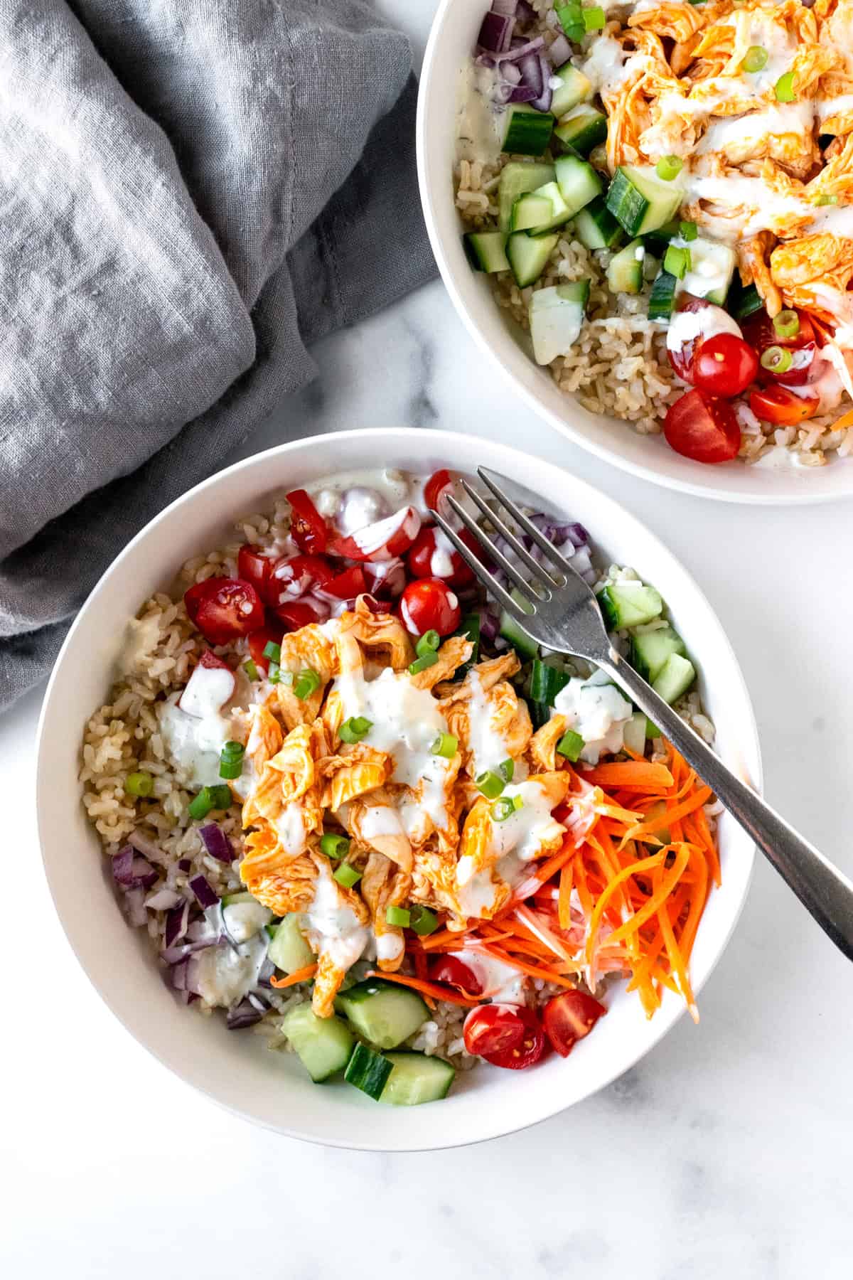Buffalo chicken and rice in two bowls topped with carrots, cucumber, tomatoes, and red onion.