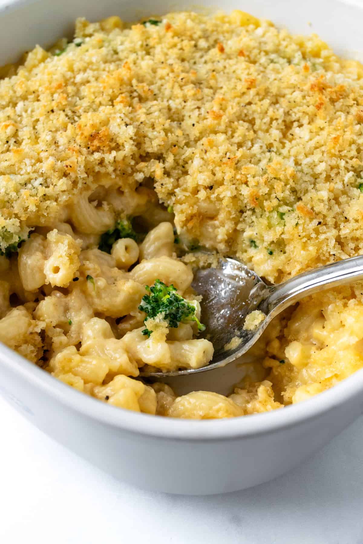 A Spoon Scooping Broccoli Mac and Cheese from a white baking dish.