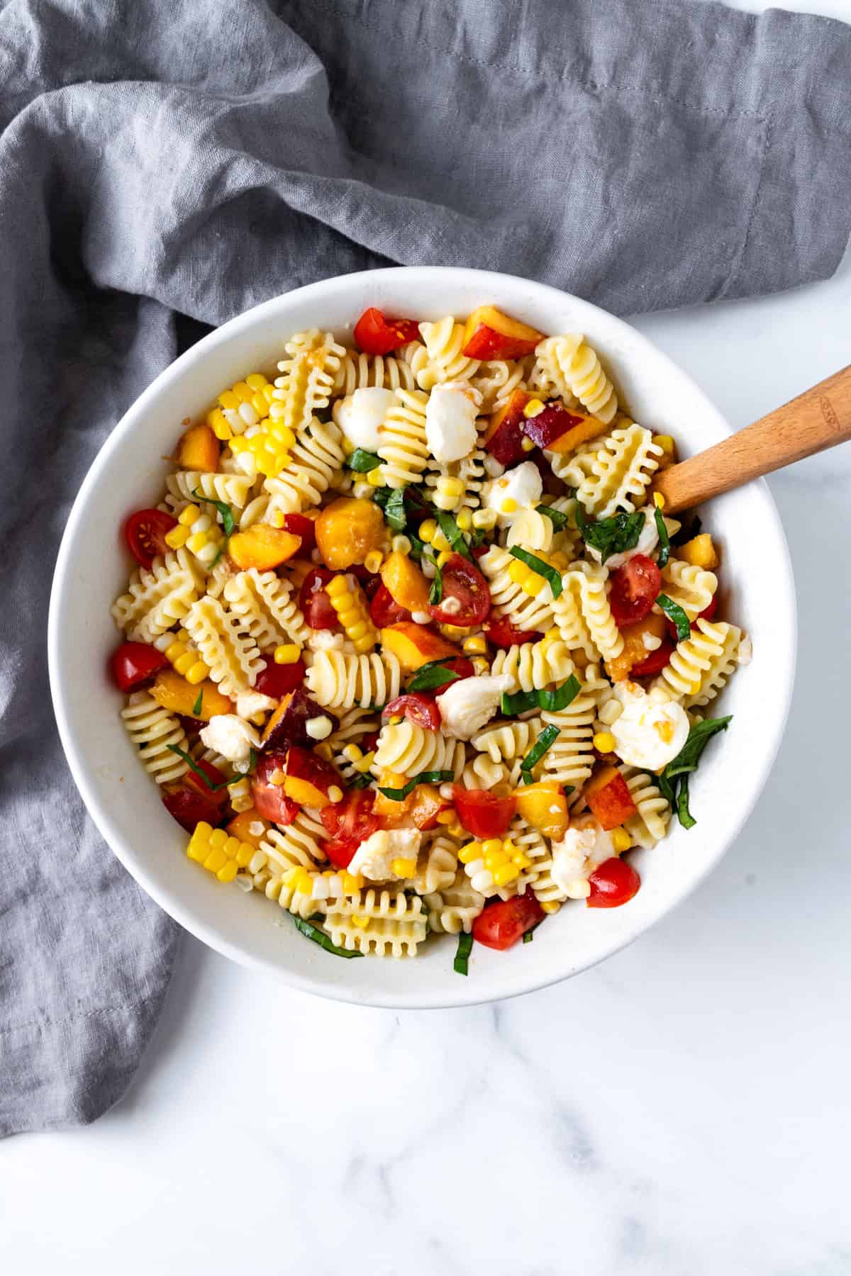 Peach and Corn Pasta Salad in a white bowl with a wooden spoon.