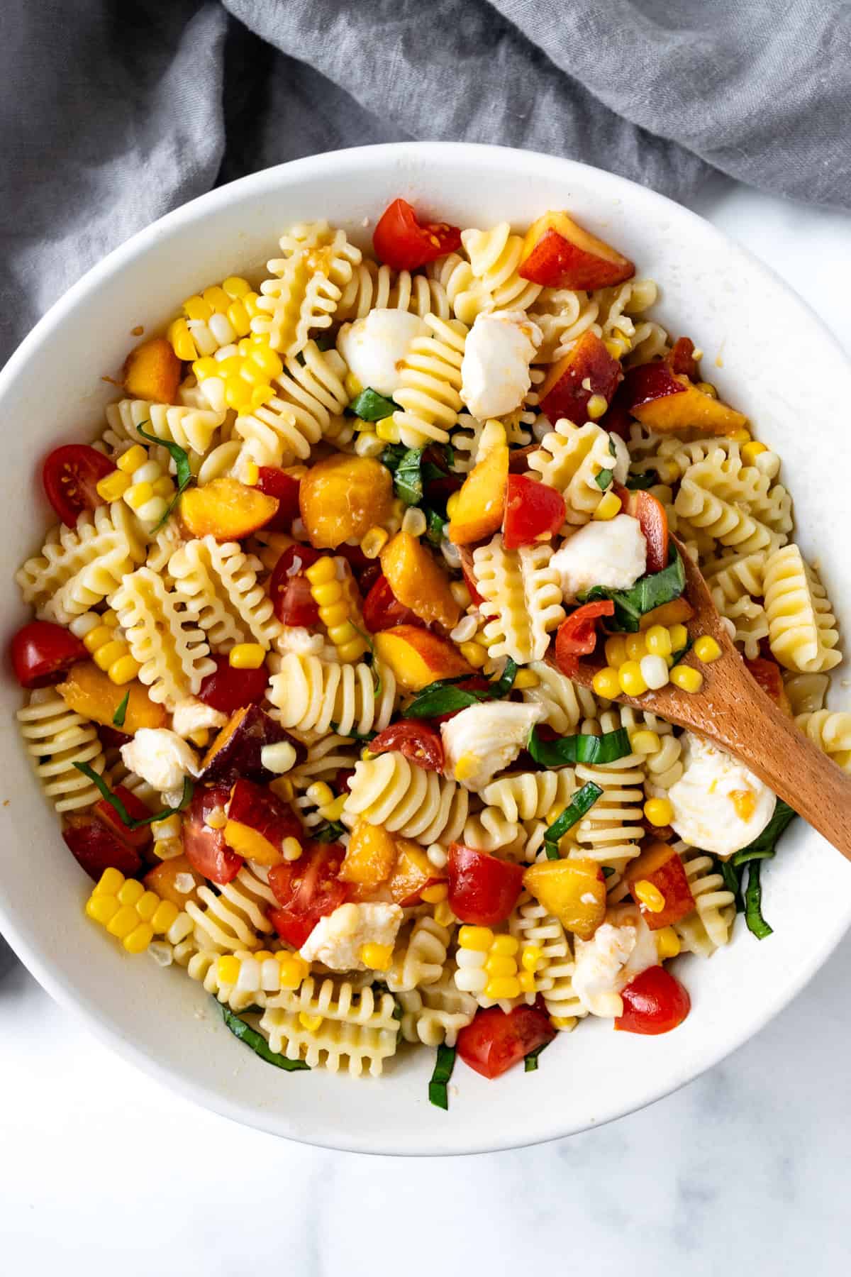 Summer Peach Pasta Salad in a white bowl with a wooden spoon.